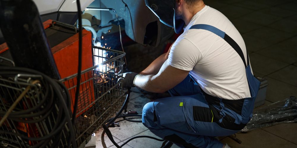 Automotive mechanic in protective helmet and gloves performing car frame welding in garage